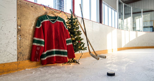 Red and green Christmas hockey jersey hanging on rink boards beside a lit mini Christmas tree, sticks and a puck