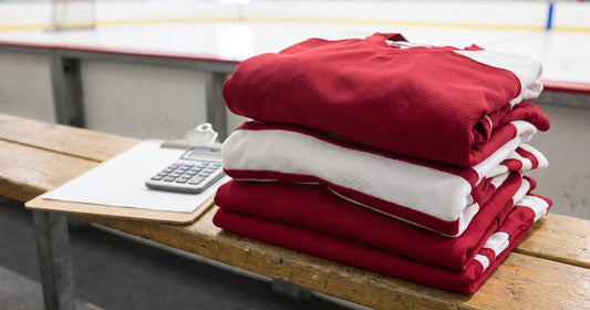 Stack of new crimson and white custom hockey jerseys on a rink bench beside a clipboard and calculator