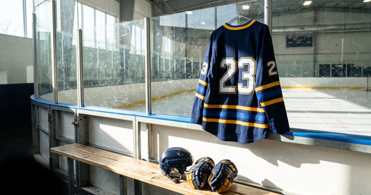 Navy and gold custom hockey jersey #23 hanging on the rink glass above a bench with a helmet and gloves