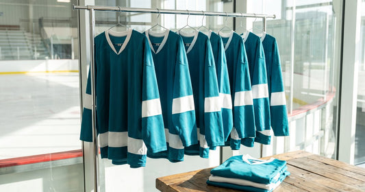Rack of matching teal and white custom hockey jerseys in a rink lobby with a folded stack on a wooden table