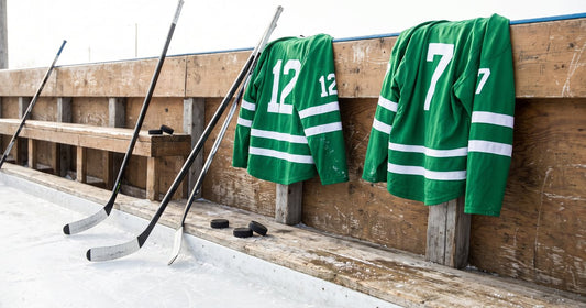 Green and white beer league jerseys hanging on the boards over the ice with taped sticks and pucks on the bench