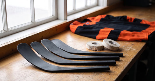 Five black hockey stick blades with different curves on a workbench beside tape rolls and an orange and black jersey