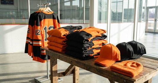 Orange and black hockey team store merch: jerseys on a rack, folded hoodies, caps and a beanie on a wooden table