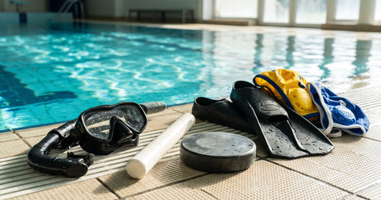 Underwater hockey gear by a pool: dive mask, snorkel, short stick, heavy puck, fins and water polo caps